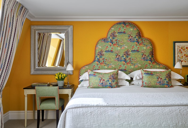 A bedroom with multi coloured headboard. There is a desk with a accent chair, and a lampshade and a mirror hanging with a flower.