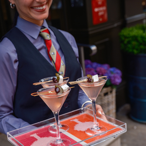 An image of a female member of Brasserie Max Bar Staff wearing uniform and holding a tray of three martinis, on a pink Ozone by Kit Kemp and Christopher Farr acrylic tray. In the street outside Brasserie Max at Covent Garden Hotel. 