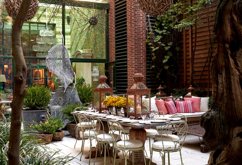 Wide shot from behind a tree, of a large stone table set up for ten guests in  the sculpture garden at the Crosby Street Hotel with lounge sofas at the back of the shot.