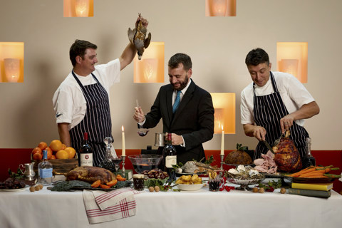 An image of a large festive table spread filled with lots of different dishes including roast ham, roast duck, and other side dishes, there are two chefs standing behind the table one is holding a bird, the other is carving the ham, the man in the middle is pouring mulled wine into a silver goblet.