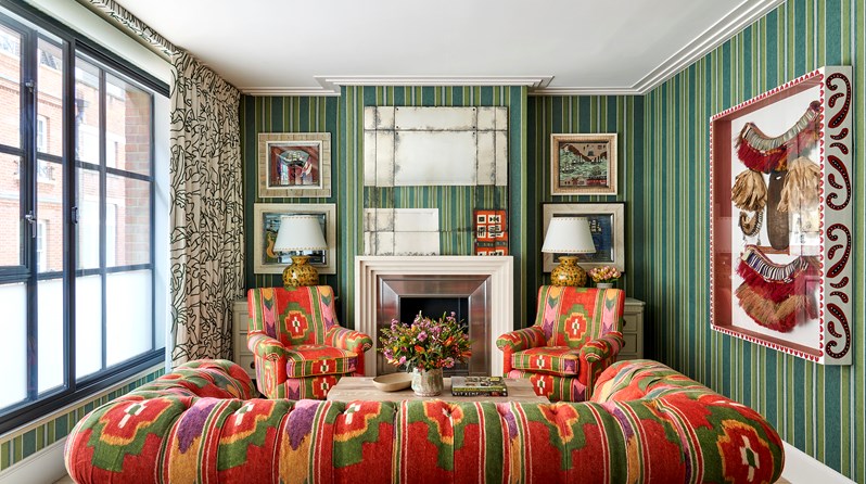 A living room space with coral red fabric walls and green and red patterned sofas and chairs. Art works hang either side of the fireplace