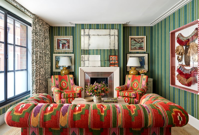 A living room space with coral red fabric walls and green and red patterned sofas and chairs. Art works hang either side of the fireplace