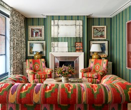 A living room space with coral red fabric walls and green and red patterned sofas and chairs. Art works hang either side of the fireplace