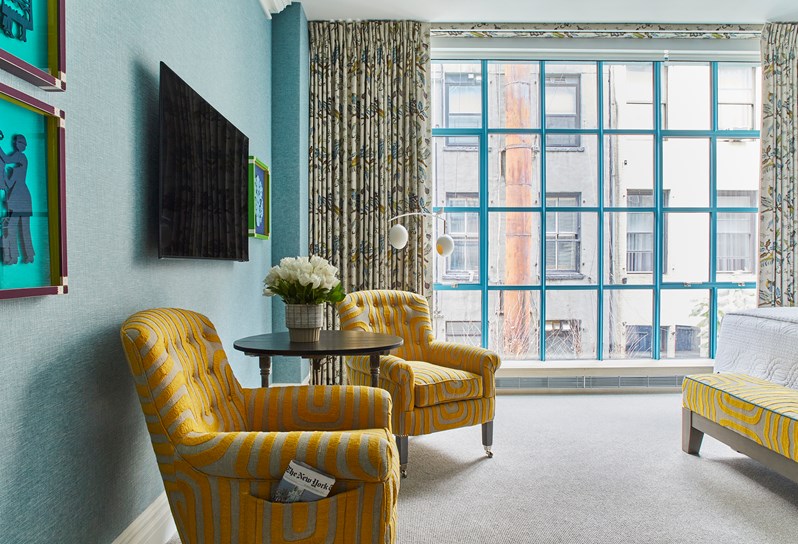 A TV and blue framed prints on a blue wall. A coffee table with white flowers and two yellow patterned armchairs with a matching ottoman
