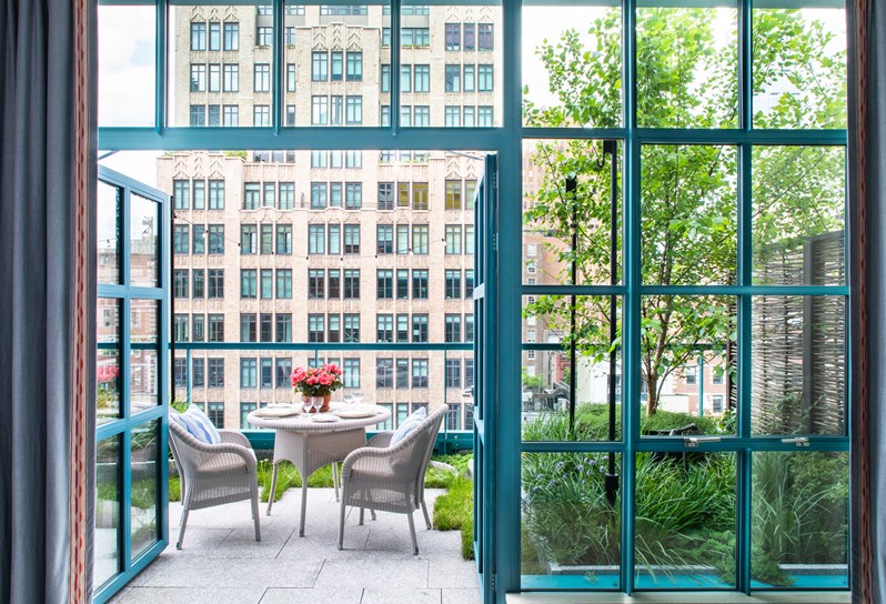 Blue windowed doors opening up onto a terrace in the city with a white wicker table and chairs decorated with blue cushions. They sit on a patio surrounded by grass and several plant pots filled with greenery.