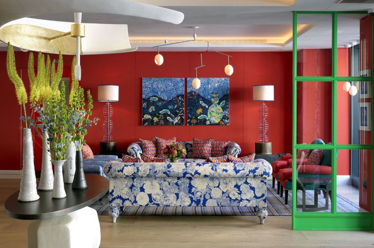 A wide shot of the lobby reception area featuring two blue and white floral sofas behind a large central sculptural table and pendant light hanging above. The walls are upholstered in a red fabric with statement artwork hung in front at 9-11 Richmond Buildings