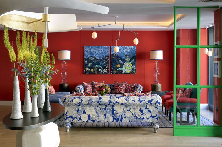 A wide shot of the lobby reception area featuring two blue and white floral sofas behind a large central sculptural table and pendant light hanging above. The walls are upholstered in a red fabric with statement artwork hung in front at 9-11 Richmond Buildings
