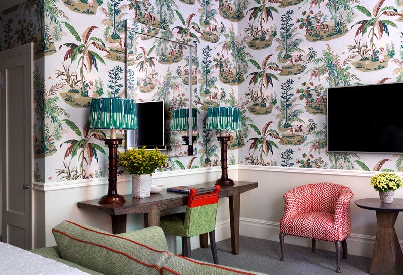 An image of the view from the bed  in Room 111, at Covent Garden Hotel. There is a desk with a green and orange chair, under a large mirror and an orange patterned chair as well as a tropical themed wallpaper.