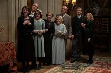Still from 'Downton Abbey: The Grand Finale'. The household staff of Downton Abbey stand in the entrance hall all looking the same way at something off screen. There are three men and 5 women all dressed all dressed in period household staff attire. 
