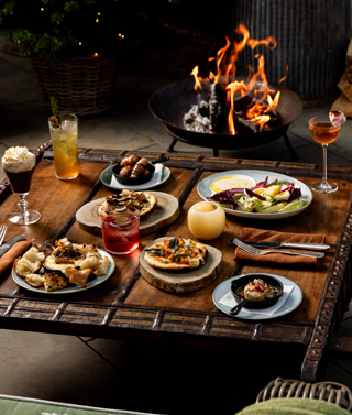 A selection of dishes and drinks on a coffee table next to a fire bowl and a tree decorated with twinkiling lights