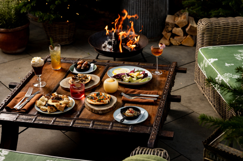 A selection of dishes and drinks on a coffee table next to a fire bowl and a tree decorated with twinkiling lights
