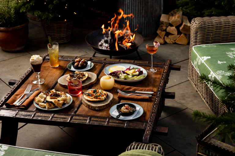 A selection of dishes and drinks on a coffee table next to a fire bowl and a tree decorated with twinkiling lights