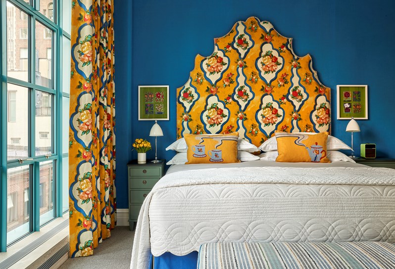 In front of blue wall, there is a bed with a yellow and blue headboard featuring a tropical fruit print which matches the curtains on the left side. There are yellow cushions with a tea cup and teapot detailing on the bed and a blue and white striped ottoman at the end. Two green floral prints hang on the wall above bedside tables either side.