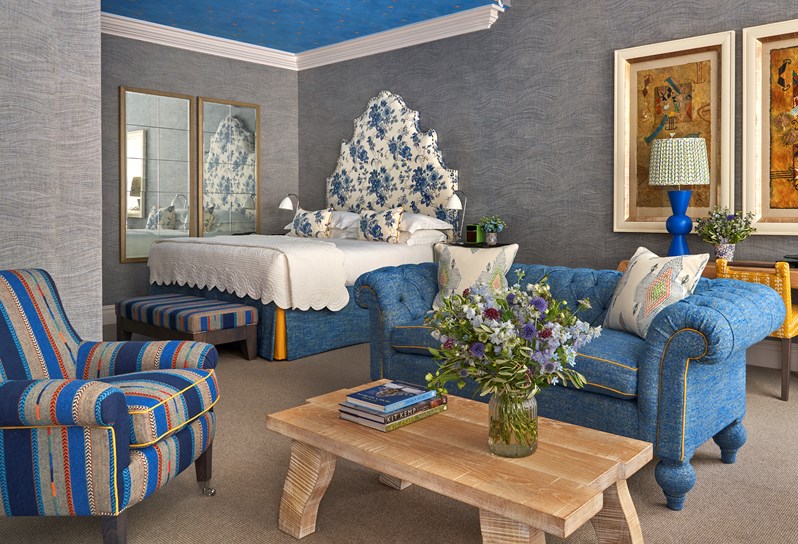 Diagonal shot of the bedroom of room 101, the Knightsbridge Suite at Knightsbridge Hotel. The image shows the seating area with blue chairs and sofa and low light wood coffee table in the foreground. in the back left of the image we see the bed with a large floral blue headboard.