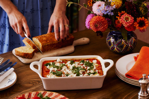 This images a landscape image that shows the final product of the recipe of Baked eggs and cornbread, here you can see a white baking dish with baked eggs in a totamto sauce topped with feta and corriander. in the bakground you can see a lady in a blue dress cutting into cornbread on a wooden board. In the foreground there is a stack of mythical creatures side plates and an orange napkin from Kit Kemp shop. 