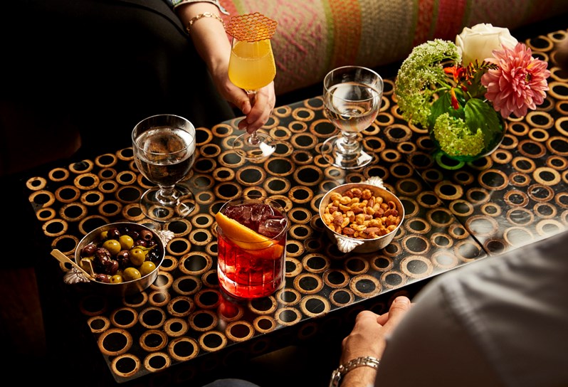 A bar table with a Negroni, a yellow-coloured cocktail in a long stemmed glass, two glasses of water, a bowl of olives and a bowl of nuts on. A man and woman can be seen partially in view, sat either side of the table.