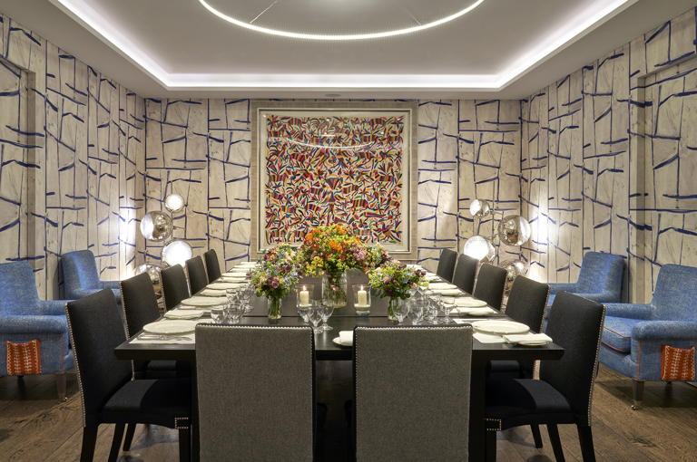 An image of the long dining table set up with flowers, place mats, plates, cutlery and glassware ready for dining in the Sandra Blow, private events room, at The Soho Hotel. With 14 grey chairs around the long tables. 