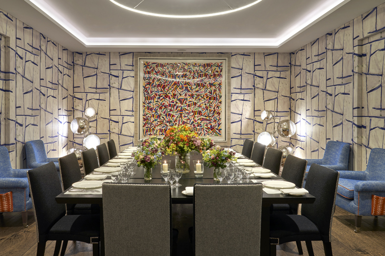 An image of the long dining table set up with flowers, place mats, plates, cutlery and glassware ready for dining in the Sandra Blow, private events room, at The Soho Hotel. With 14 grey chairs around the long tables.