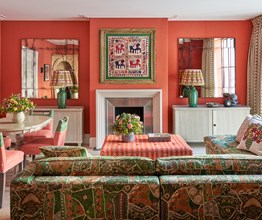 A living room space of the Deluxe Two Bedroom Suite Room 5 at Ham Yard Hotel.  with red-orange walls, patterned green furniture, and eclectic decor including mirrors, floral arrangements, and framed textile art above the fireplace. The image shows the back of a 'L shaped' sofa on the right hand side, pointing towards the fire place. On the left the dining table with 4 chairs around