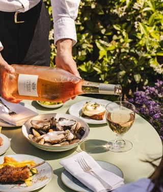 An image of a someone pouring a bottle Chateau Le Coste with some dishes from the 'Rose on the Roof' menu at Ham Yard Hotel.