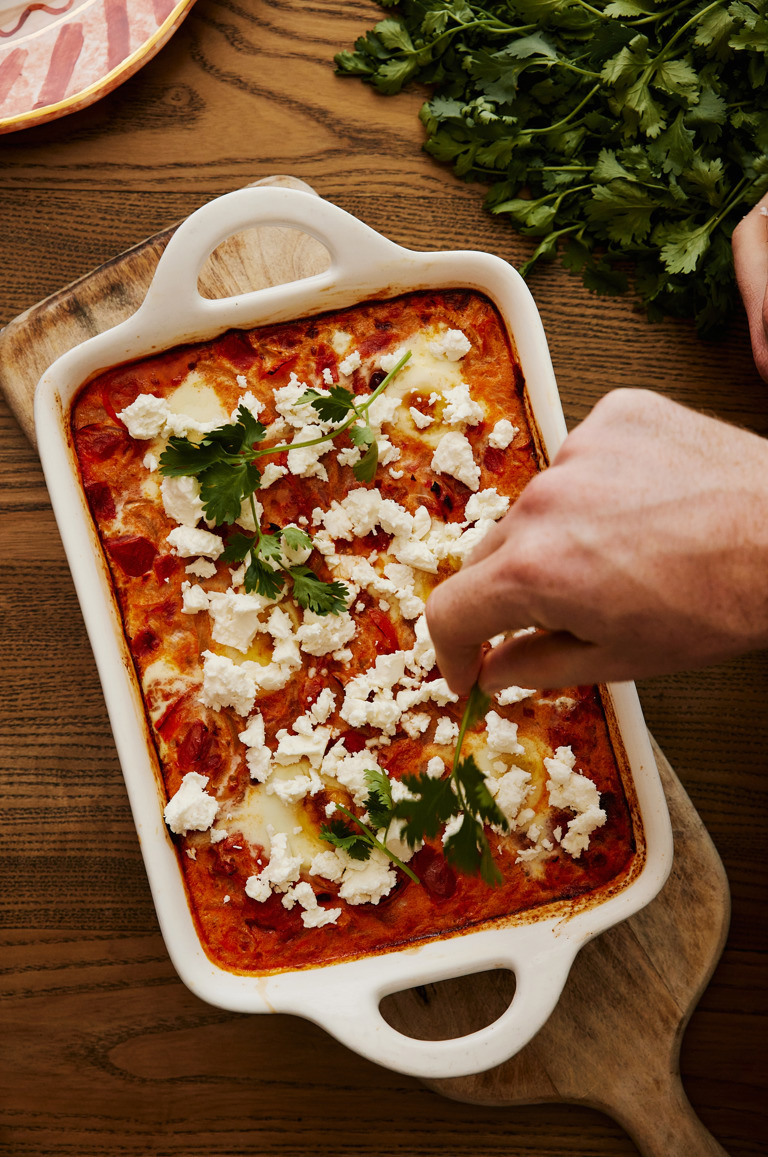 In this image you can see the final product of the baked eggs fresh from the oven with a hand sprinkling feta and coriander on top.  