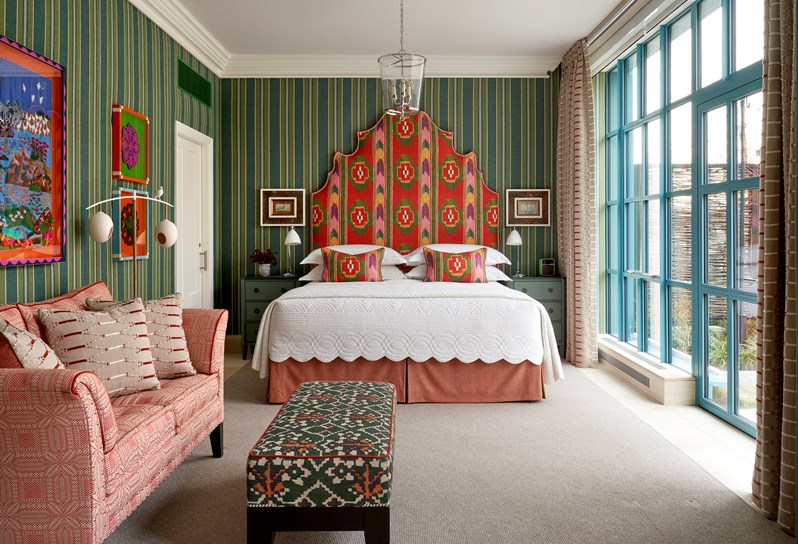 A bedroom with green striped walls and floor to ceiling windows and doors that open out onto a terrace. A bed with a multi-coloured geometric print headboard and matching cushions. On the left, a pink patterned sofa and a green and orange patterned footstall.