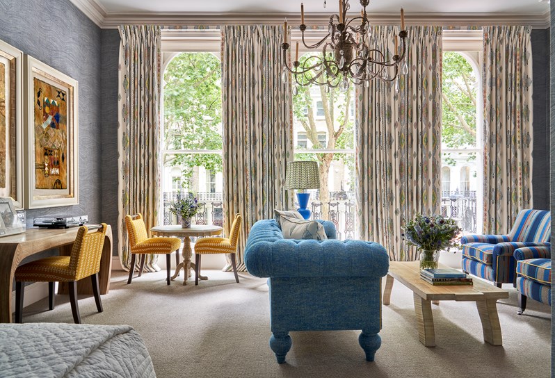 Living room shot of room 101, the Knightsbridge Suite at Knightsbridge Hotel. The image shows a blue sofa from the side in the centre of the image facing towards two blue striped arm chairs and a coffee table. in the left we see two yellow chairs round a small circular tale and a yellow chair in front of a desk.
