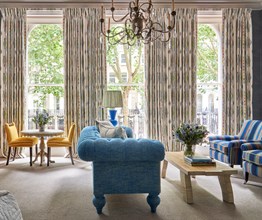 Living room shot of room 101, the Knightsbridge Suite at Knightsbridge Hotel. The image shows a blue sofa from the side in the centre of the image facing towards two blue striped arm chairs and a coffee table. in the left we see two yellow chairs round a small circular tale and a yellow chair in front of a desk.