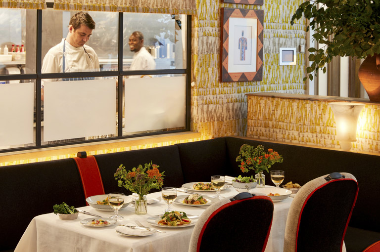 An image to promote the Summer à la carte menu at Ham Yard Bar & Restaurant at Ham Yard Hotel. The image shows a corner table set with white linen, small floral arrangements, and plates of freshly prepared dishes. Glasses of white wine are placed alongside the plates, and chefs are visible working in the open kitchen behind.