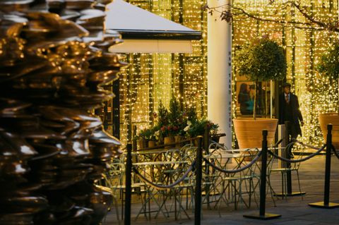A diagonal shot looking from the corner of Ham Yard Hotel courtyard to the hotel entrance. A bronze sculpture by Tony Cragg can be seen close up on the left. Groups of outdoor dining tables and chairs are set up by the hotel entrance which is dressed in twinkling lights. A doorman is seen walking out of the door