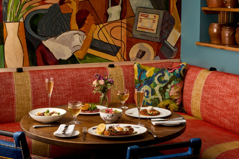 An image to promote Oscar Restaurant’s À La Carte menu at Charlotte Street Hotel. The image shows a round dark wood table with multiple dishes, examples of dishes include chicken schnitzel and trout dishes and with flutes of champagne and glasses of wine on the table.