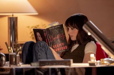 A still image from the film Verity this image shows Dakota Johnson sitting in a chair reading a book with a picture of Anne Hathaway on it.