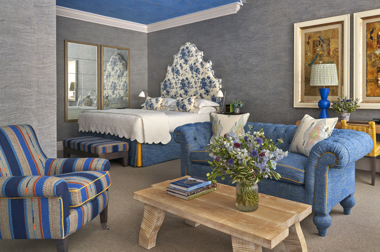 Diagonal shot of the bedroom of room 101, the Knightsbridge Suite at Knightsbridge Hotel. The image shows the seating area with blue chairs and sofa and low light wood coffee table in the foreground. in the back left of the image we see the bed with a large floral blue headboard. 