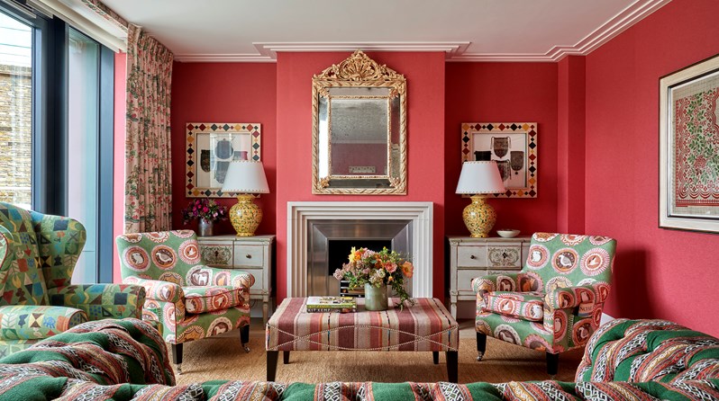 A living room space with coral red fabric walls and green and red patterned sofas and chairs. Art works hang either side of the fireplace and bi-fold doors open onto a terrace