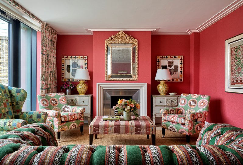 A living room space with coral red fabric walls and green and red patterned sofas and chairs. Art works hang either side of the fireplace and bi-fold doors open onto a terrace