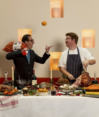 A portrait image of a large festive table spread filled with lots of different dishes including roast ham, roast duck, and a large Christmas pudding, bottle of wine and mulled wine, behind the table a sommelier throws and orange in the air as a chef carves the ham.  