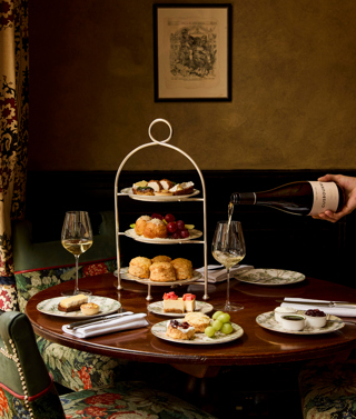 A portrait shot of the items in Afternoon Cheese at Brasserie Max at Covent Garden Hotel. On the table, there is a stand holding plates of scones and cheese-inspired pastries, and a bottle of white wine is being poured.