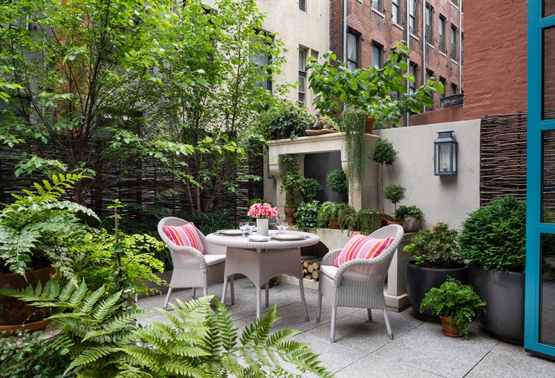 A terrace in the city with a white wicker table and chairs decorated with coral cushions. in the foreground, there are several plant pots filled with greenery and at the back of the terrace there is a stone fireplace decorated with plants