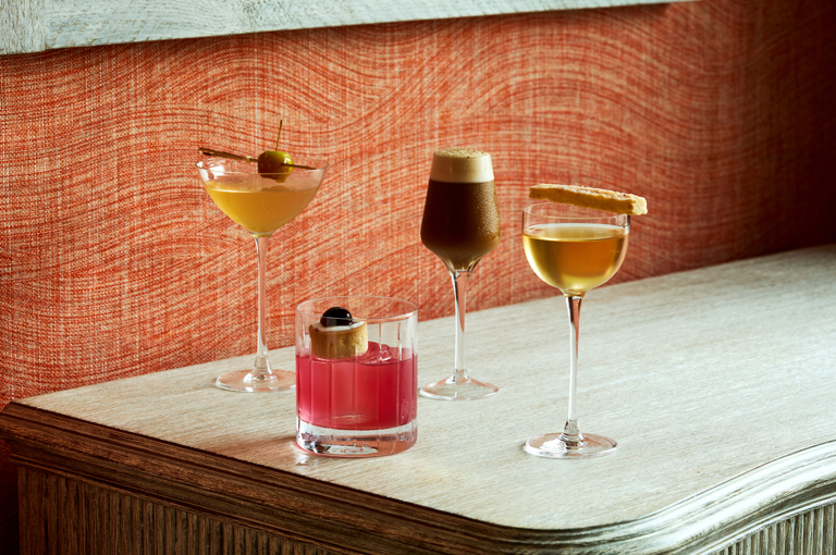 An image of four cocktails , three tall martini shaped glasses and one short whisky glass, against a fabric orange backdrop. 