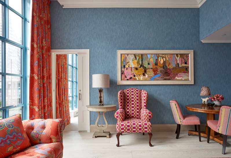 A living area. A corner of an orange 'Chainstitch' sofa which matches curtains on the windows behind. A pink and white checkered armchair and a side table with a lamp on next to a door opening into the bedroom. A round table with two red, yellow and blue chairs around it.