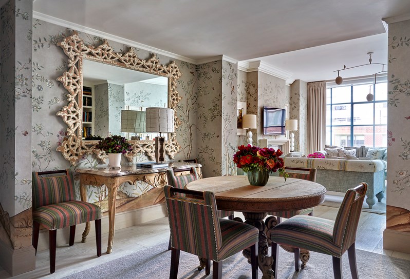 Flowers on a  round wooden table surrounded by 4 multi-coloured striped chairs. A striped chair, and marble topped console table with a lamp and flowers on in front of a mirror.