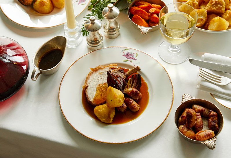 A closeup of a plate of festive roast being served on a Mythical Creatures plate. The roast includes Pork chops with gravy, pigs in a blanket, roast potatoes and roast onions. There is a glass of white wine being paired.