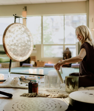 Sound practitioner Nancy Truman leading a sound bath class. She is sat on the floor playing with two glass sound bowls. In front of her are two large gongs