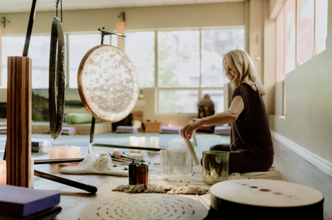 Sound practitioner Nancy Truman leading a sound bath class. She is sat on the floor playing with two glass sound bowls. In front of her are two large gongs
