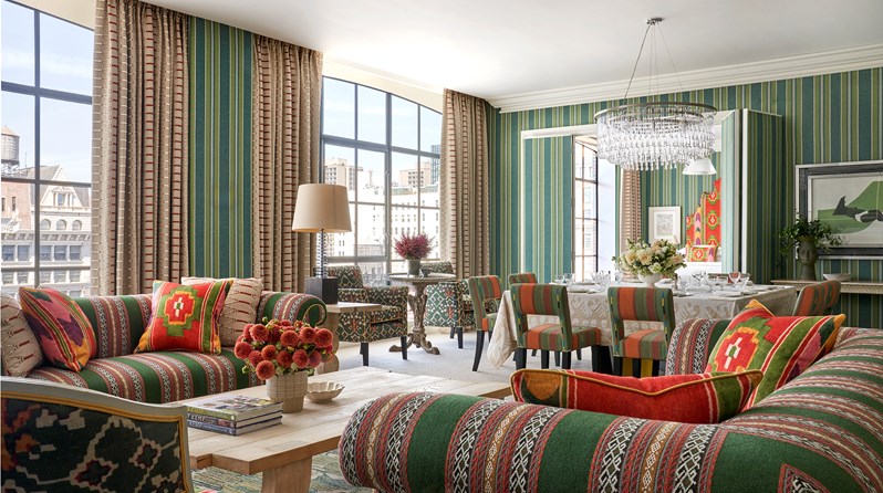 Diagonal angle shot of the living room in a One Bedroom Suite at the Crosby Street Hotel with two sofas around a coffee table in first plan, large windows on the back left and a dinner table set up with 6 chairs on the back right just in front of open doors leading to the bedroom.
