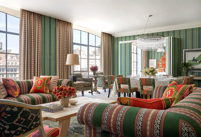 Diagonal angle shot of the living room in a One Bedroom Suite at the Crosby Street Hotel with two sofas around a coffee table in first plan, large windows on the back left and a dinner table set up with 6 chairs on the back right just in front of open doors leading to the bedroom.