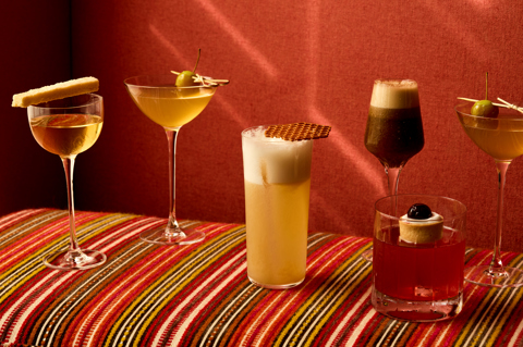 Image of 6 cocktails perched on stripy furniture in front of a red fabric wall. 3 in tall glasses, one tall glass and one short whisky glass.