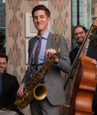 The Sam Taylor Jazz Trio. One man stands holding a saxophone in the middle, to the left there is man sat behind a drum and to the ridge, there is a man holding a bass