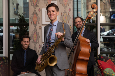 The Sam Taylor Jazz Trio. One man stands holding a saxophone in the middle, to the left there is man sat behind a drum and to the ridge, there is a man holding a bass