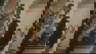 Still from 'The Mastermind'. A bearded man dressed smartly, stands in front of an oil painting in an art gallery.